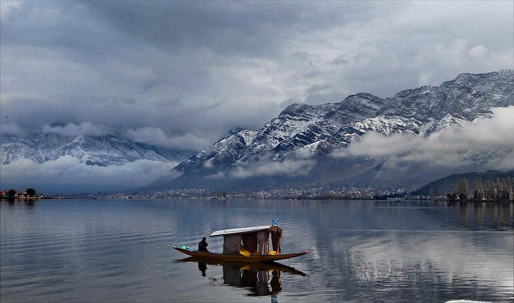 A shikarawala rowing his boat on Dal Lake with snow covered mountains in the background, in Srinagar.