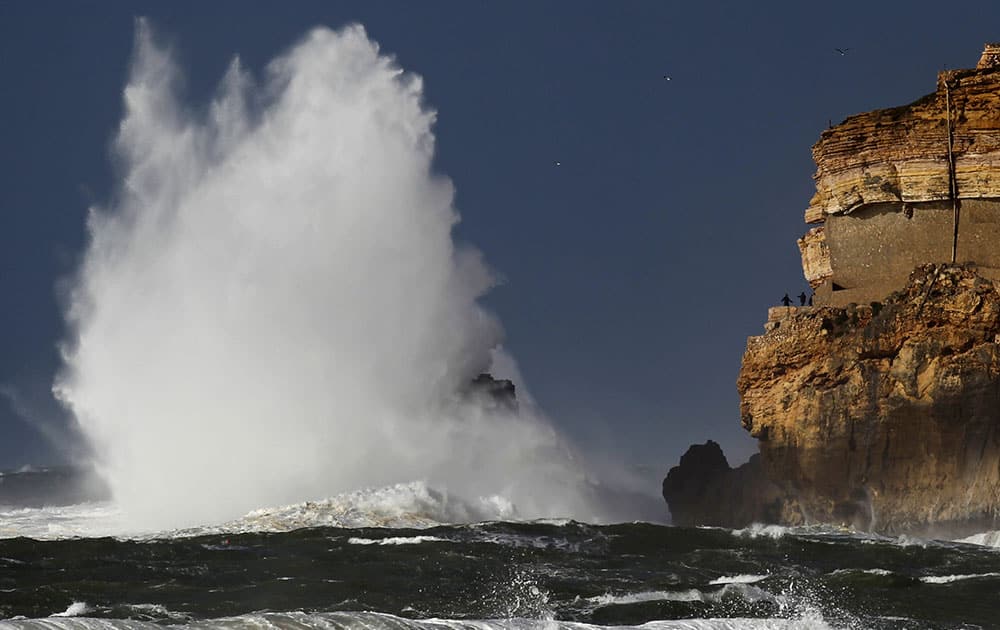 People, in a viewpoint of a cliffside, look at waves breaking against rocks at the Nazare lighthouse, outside the fishing village of Nazare, central coast of Portugal.