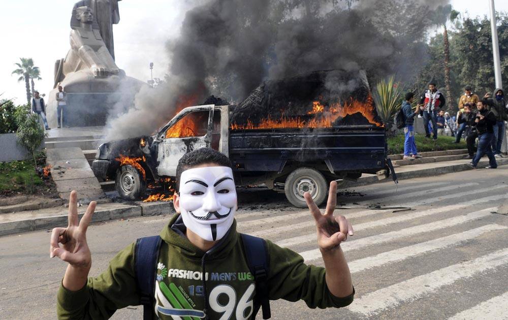 A protester wearing a Guy Fawkes mask gives the victory sign in front of a burning police vehicle set afire by protesters, near Cairo University, Cairo.