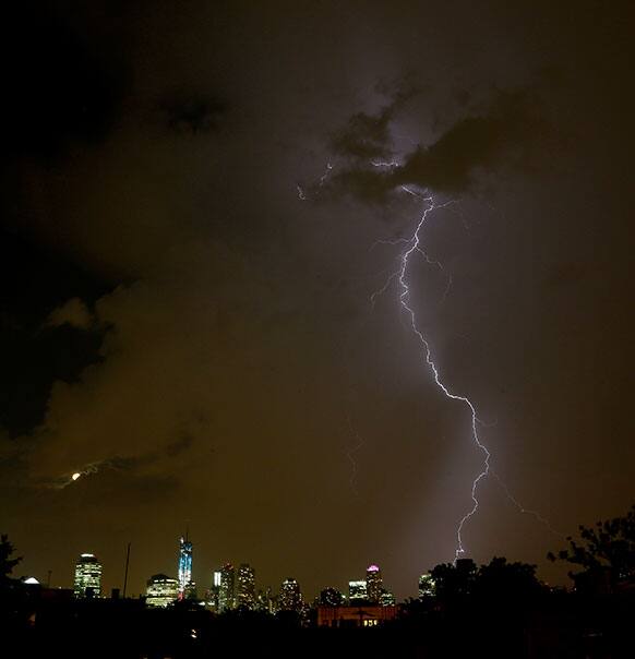 With One World Trade Center, dressed in red, white and blue lights and a moon in its waning gibbous stage peeking through clouds, left, a lightning bolt from a thunderstorm strikes down over Lower Manhattan as seen from The Heights neighborhood of Jersey City, N.J.