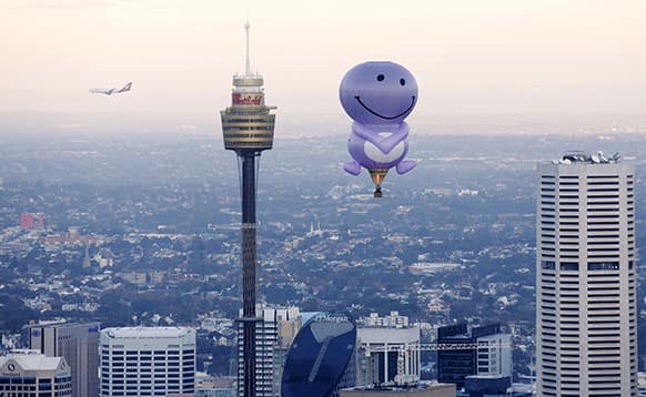 A hot air balloon from a fruit juice company flies over Sydney Harbour and along the Sydney skyline as a commercial jet flies past in the back.