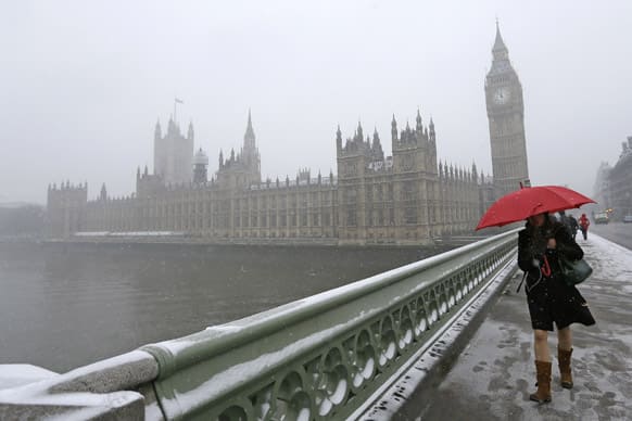 A woman carrying a red umbrella walks across Westminster Bridge, with the Palace of Westminster in the background as it begins to snow in London.