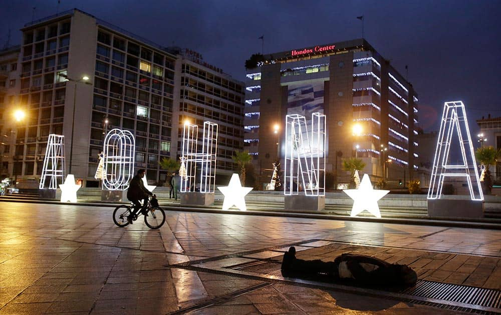 A man rides his bicycle next to a lit Christmas sign reading ''Athens'' as a homeless man sleeps on a metro air vent above an underground station to warm up, at Omonia Square in central Athens.