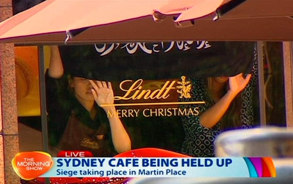 This image taken from video shows people holding up what appeared to be a black flag with white Arabic writing on it, inside a cafe in Sydney, Australia. An apparent hostage situation was unfolding inside the chocolate shop and cafe in Australia's largest city on Monday, where several people could be seen through a window with their hands held in the air.