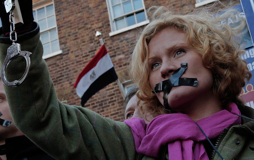 A journalist, with black tape over the mouth to illustrate the silencing of free speech and journalists, holds up a placard and a pair of handcuffs as she demonstrates across the street from the Egyptian Embassy to mark a year since the detention of Al-Jazeera English journalists in Egypt, central London.