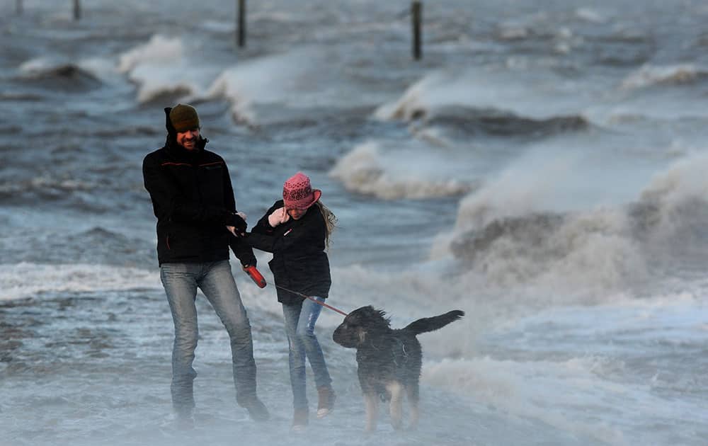 A father with his daughter and a dog make their way through string winds as big storm waves pound on the flooded quay at the North Sea in Neuharlingersiel, northern Germany, Strong winds have caused disruption in Scotland and northern Germany, with several railway lines closed. National railway Deutsche Bahn said routes from Hamburg to Hannover, Berlin and Bremen were closed on Friday, along with several others in the country's north, due to uprooted trees and high winds.
