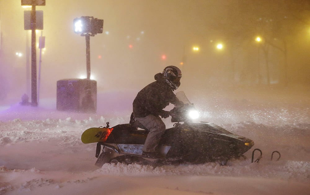 A man uses a snowmobile to travel along a street before dawn during a winter snowstorm.