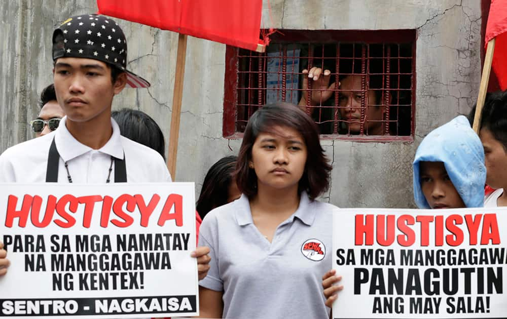 A private guard looks from a small window as protesters display placards during a rally outside the burnt Kentex rubber slipper factory in Valenzuela city, a northern suburb of Manila, Philippines.