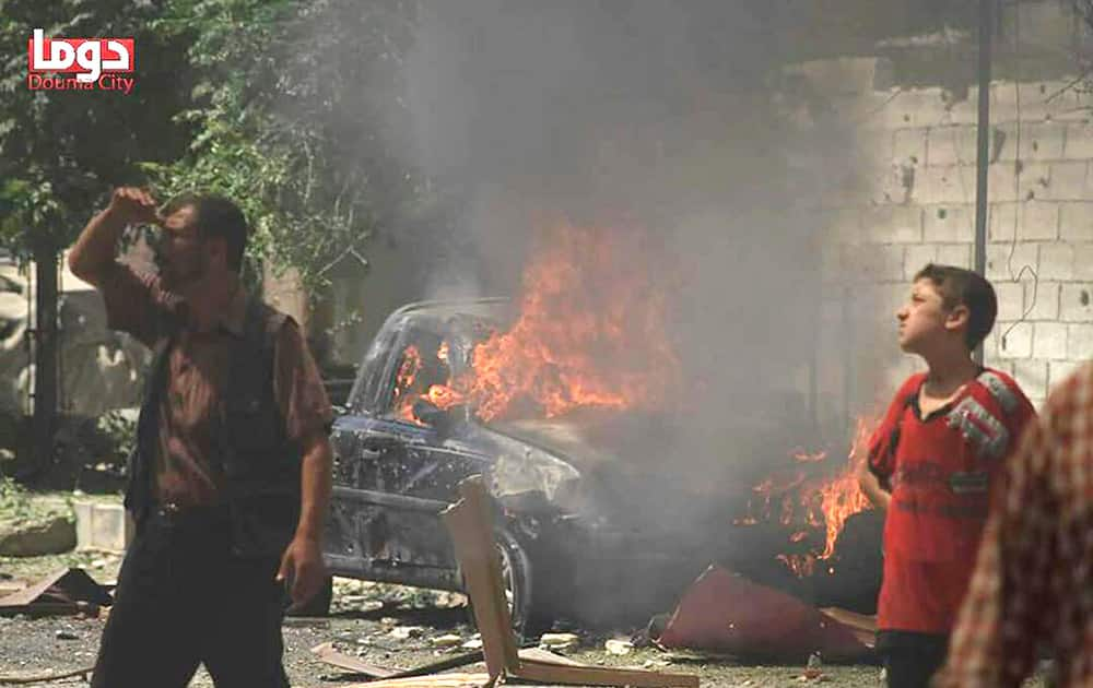 Syrian citizens look on as they stand in front of a burned car that set on fire by an attack of Syrian government airstrike, in Douma, a suburb of Damascus, Syria.