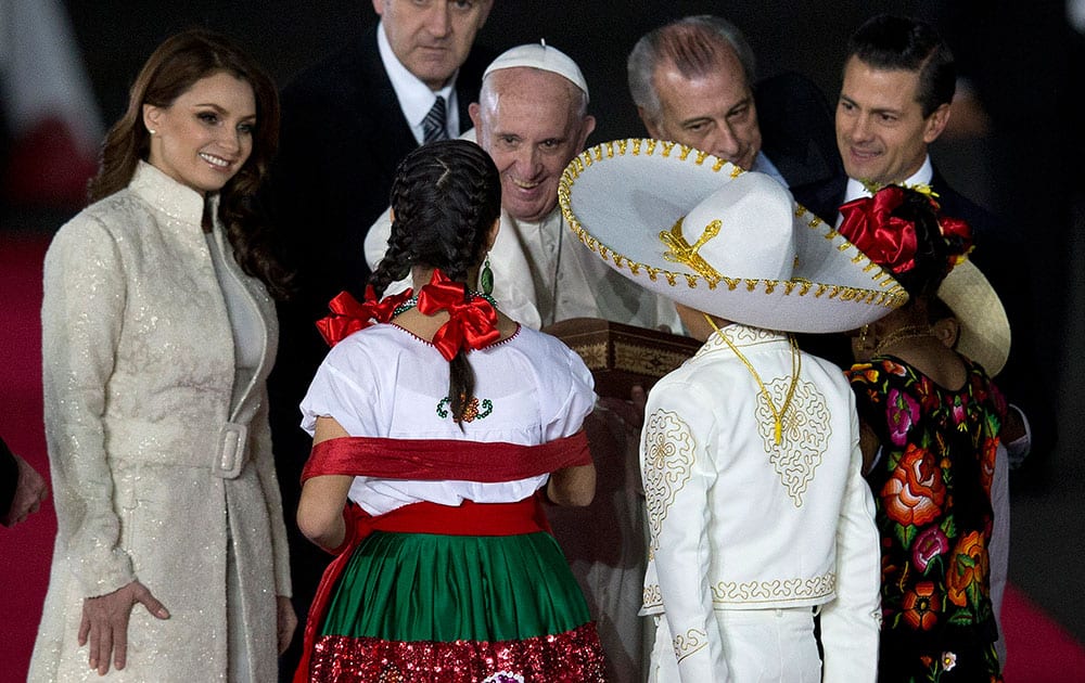Pope Francis reaches out to greet youth dressed in traditional Mexican outfits as he's escorted by Mexico's President Enrique Pena Nieto, behind right, and first lady Angelica Rivera upon arrival to Benito Juarez International Airport in Mexico City.