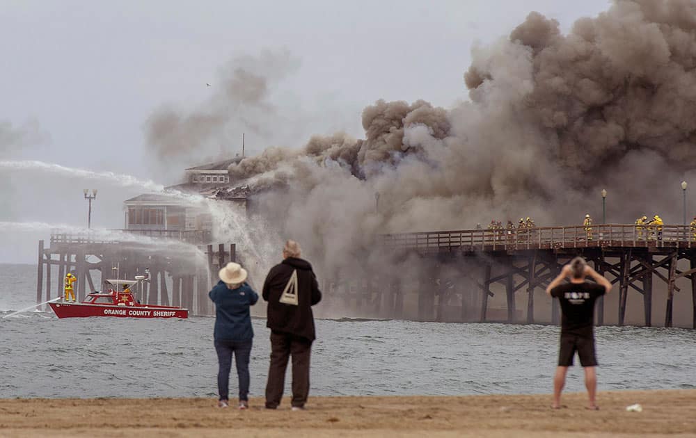 Spectators gather on the sand as fire boats and firefighters battle a fire on the Seal Beach pier in Seal Beach, Calif.