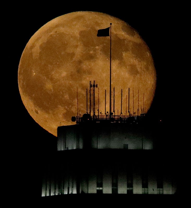 A waning full moon rises beyond an American flag flying over City Hall