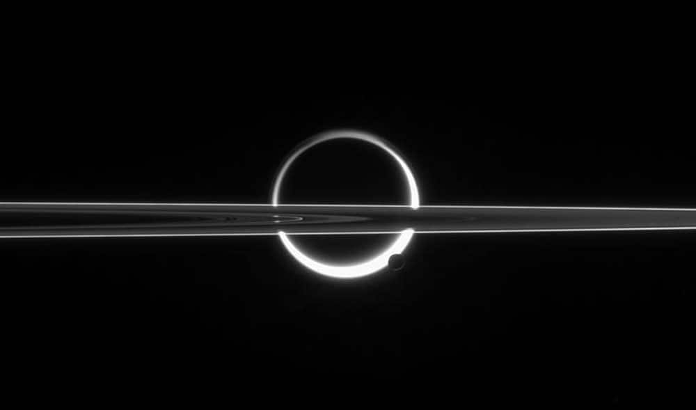 Saturn's rings cut across an eerie scene that is ruled by Titan's luminous crescent and globe-encircling haze, broken by the small moon Enceladus, whose icy jets are dimly visible at its south pole