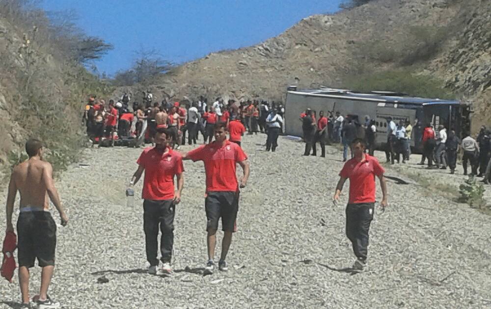 Emergency personnel and members of Agentina's Huracan soccer team, in red shirts and black shorts, mill around the wreck of the bus that was transporting the team to the airport, after it lost its brakes, lost control and flipped over, in Caracas.